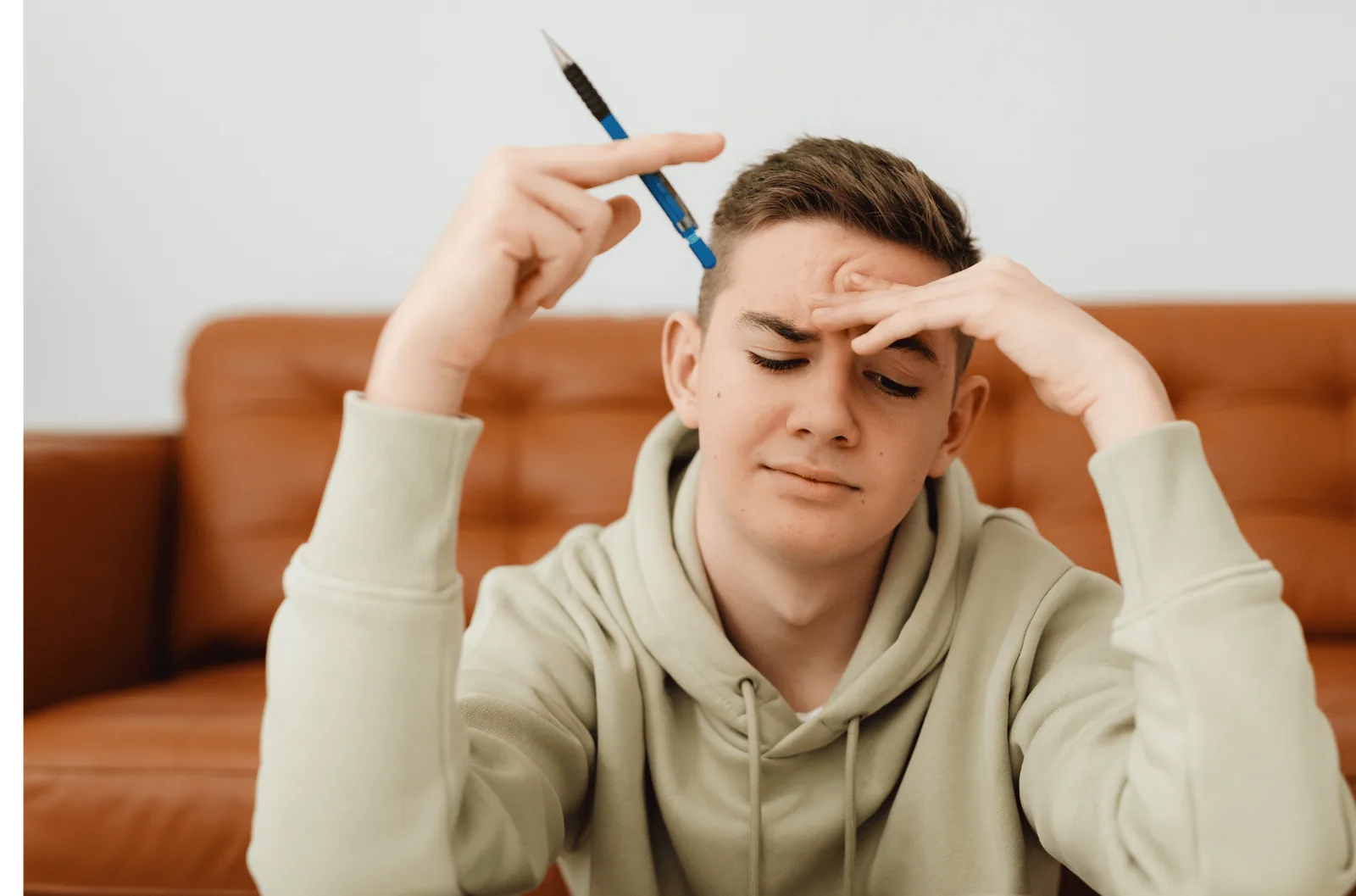 Young person sitting on a sofa with a pencil and hands against their forehead.